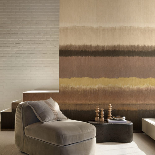 A spacious interior featuring the horizontally striped Pianura wallcovering by Omexco, a vision of misty landscapes, animated by rich tonal nuances. The design flows with earthy bands of brown, beige, and mustard next to a white painted brick wall, framing a cozy area with a beige armchair.