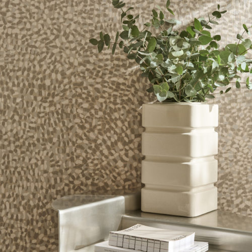 Gros plan du revêtement mural Pianura beige clair et gris d'Omexco, affichant des accents de pelage, qui anime les murs avec caractère. Le délicat motif tacheté forme une toile de fond pour un vase blanc carré nervuré contenant des branches d'eucalyptus vert sur une table métallique.