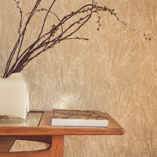 Nahaufnahme der goldbeigen Whisper Wandbekleidung von Omexco mit einem stilisierten Motiv, inspiriert von feinen mineralischen Strukturen und Muscheln. Die wunderschön strukturierte Wand befindet sich hinter einem Holztisch im Mid-Century-Stil mit einer weißen Vase und dekorativen Zweigen.