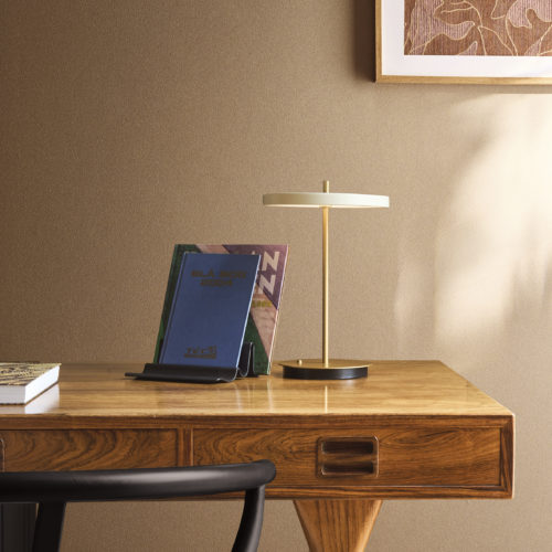 Ein Home-Office-Bereich mit der braunen Whisper Wandbekleidung von Omexco, bei der sanft aufgestäubtes Reishülsenpulver ein filigranes Muster mineralischer Aderungen zeichnet. Die warm strukturierte Wand passt hervorragend zu einem Holzschreibtisch, einer modernen Messinglampe und einem gerahmten abstrakten Kunstwerk.
