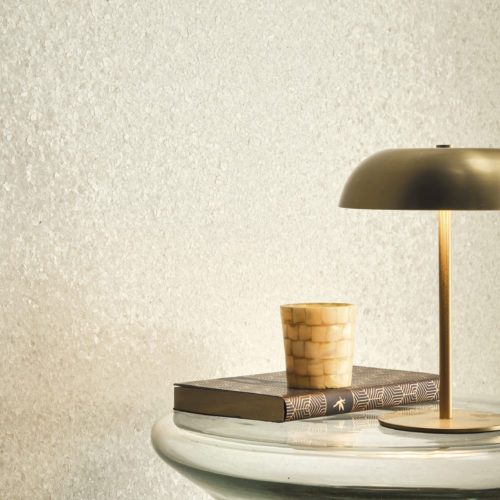Interieurdetail, das die funkelnd weiße Whisper Wandbekleidung von Omexco hervorhebt, wunderschön strukturiert mit zarten Muschelfragmenten, die einen sanften, perlmuttartigen Schimmer erzeugen. Die leuchtende Wand schafft eine ruhige Atmosphäre hinter einem skulpturalen Glastisch, der mit einer modernen Messinglampe und einem Mosaik-Kerzenhalter verziert ist.