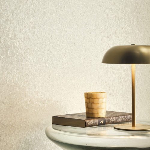 Interieurdetail, das die funkelnd weiße Whisper Wandbekleidung von Omexco hervorhebt, wunderschön strukturiert mit zarten Muschelfragmenten, die einen sanften, perlmuttartigen Schimmer erzeugen. Die leuchtende Wand schafft eine ruhige Atmosphäre hinter einem skulpturalen Glastisch, der mit einer modernen Messinglampe und einem Mosaik-Kerzenhalter verziert ist.