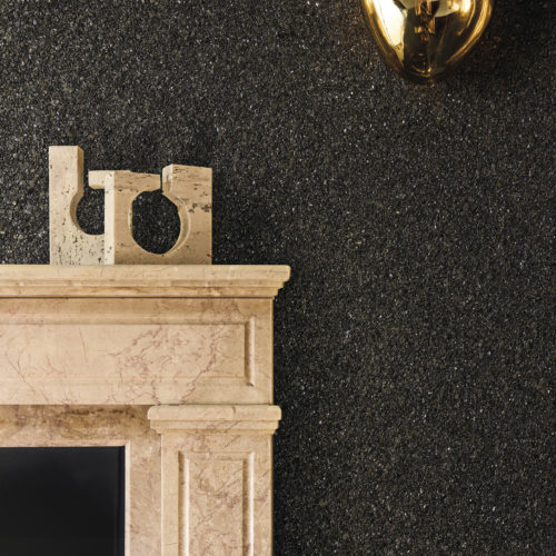 Eleganter Raum mit der dunklen Whisper Wandbekleidung von Omexco aus strukturiertem Mica in einer neuen Farbpalette. Die funkelnde schwarze Wand bildet einen schönen Kontrast zu einem klassischen Kamin aus rosafarbenem Marmor und einer modernen bernsteinfarbenen Wandleuchte.