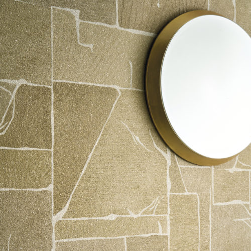 Detail der goldenen Whisper Wandbekleidung von Omexco, die Intarsien in Marmoroptik mit fein ausgearbeiteten Details auf einem Mica-Hintergrund präsentiert. Die geometrische, strukturierte Wand ist elegant mit einem schlichten runden Spiegel mit Messingrahmen verziert.