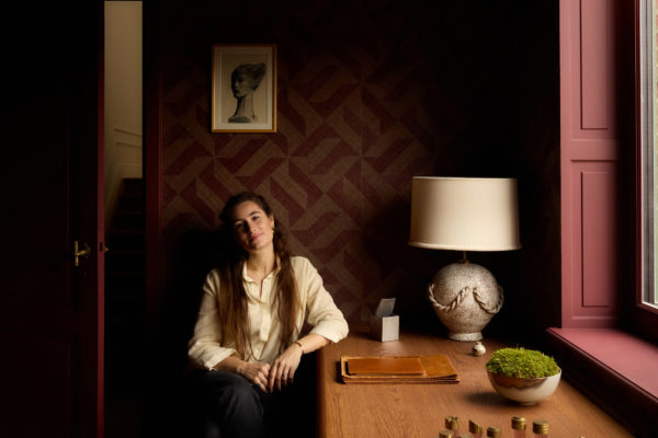 Interior portrait of interior architect Aurélie Penneman seated at a wooden desk in a historic Ghent townhouse. A geometric wallcovering in deep red and natural tones forms a textured backdrop, complemented by sculptural lighting, curated objects and a warm, contemporary atmosphere.