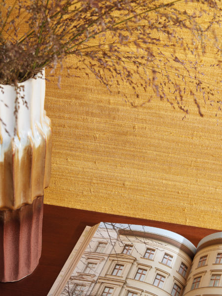 Gros plan d'un livre d'architecture ouvert posé à côté d'un vase en céramique tricolore des années 70. Le vase contient des fleurs séchées. Les deux objets sont posés sur une table ou un buffet en bois, contre un mur ocre orné d'un précieux motif textile en soie. Il s'agit des revêtements muraux "Loom Stories" d'Omexco, une collection riche et éclectique de textures irrégulières réalisées à partir de fils filés à la main, de lin belge fin et de soie sauvage, le tout dans une palette variée allant de tons neutres apaisants à des couleurs vives et audacieuses qui attirent l'attention.