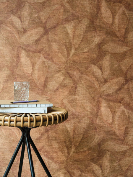 Cet intérieur accueillant met en valeur la référence OAS202, Hierbas, issue de la collection Oase d’Omexco. Le revêtement mural présente un motif délicat de feuilles imprimé sur du raphia naturel, créant une atmosphère chaleureuse et organique. Les tons terreux de terracotta du mur s’accordent parfaitement avec la table d’appoint en rotin et métal, apportant une touche à la fois rustique et raffinée. Une pile de livres, un verre d’eau et un stylo posés sur la table renforcent l’ambiance détendue et sophistiquée, idéale pour des moments de calme et de réflexion.