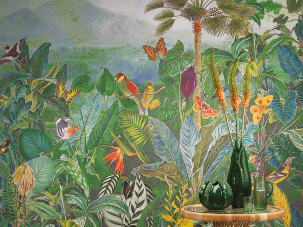 Gros plan d'une grande impression murale de jungle montrant des plantes exotiques, des oiseaux du paradis, des palmiers et des papillons. Ce décor panoramique à quatre gouttes de la collection "Birds of Paradise" d'Omexco est imprimé sur des revêtements muraux intissés et nous plonge dans la splendeur de la nature. Devant le mur, nous voyons un pouf rond ocre ainsi qu'une table d'appoint ratan sur laquelle sont posés deux vases en céramique verte.