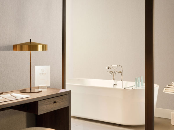 Softly textured in beige, Omexco's High Performance Essence wallpaper sets a tranquil tone, paired with a dark wood desk, golden lamp, and a glimpse of a spa-like bathroom with freestanding tub.