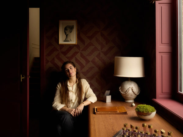 Interior portrait of interior architect Aurélie Penneman seated at a wooden desk in a historic Ghent townhouse. A geometric wallcovering in deep red and natural tones forms a textured backdrop, complemented by sculptural lighting, curated objects and a warm, contemporary atmosphere.