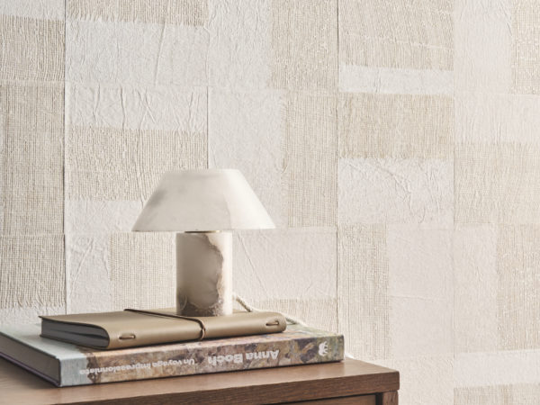 A refined interior featuring Omexco's Pure collection, reference PU261, a wallcovering named Intensify, showcasing a geometric paper patchwork enriched with linen-toned openwork abaca weaving. This design adds depth, texture, and a tactile dimension to the space, creating a striking visual statement. The scene includes a minimalist wooden cabinet with a subtle brass handle, topped with a stack of books and a leather notebook, reflecting thoughtful organization. A marble table lamp with a sculptural quality enhances the luxurious yet understated aesthetic. The harmonious combination of organic textures and geometric patterns makes this wallpaper a focal point for modern, sophisticated interiors.