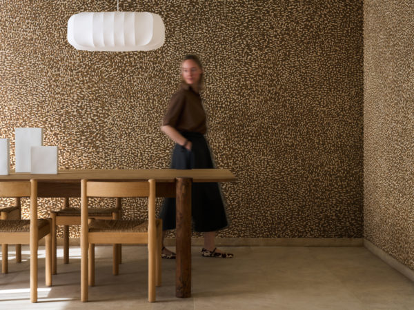 A person walking past the patterned Pianura wallcovering by Omexco with echoes of animal coats, bringing walls to life with instinct and movement. The vibrant brown and white speckled wall wraps around the corner of a dining room featuring a robust wooden table and chairs.