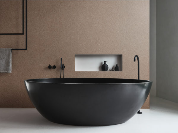 A luxurious bathroom featuring a striking black freestanding oval-shaped bathtub with modern taps and accessories. The walls are adorned with Omexco's high-performance wallcovering collection, Rivage, boasting textured tonal stripes in a captivating brown/red hue. The design adds a touch of sophistication and warmth to the space. Behind the bathtub, a niche in the wall provides a stylish storage solution.