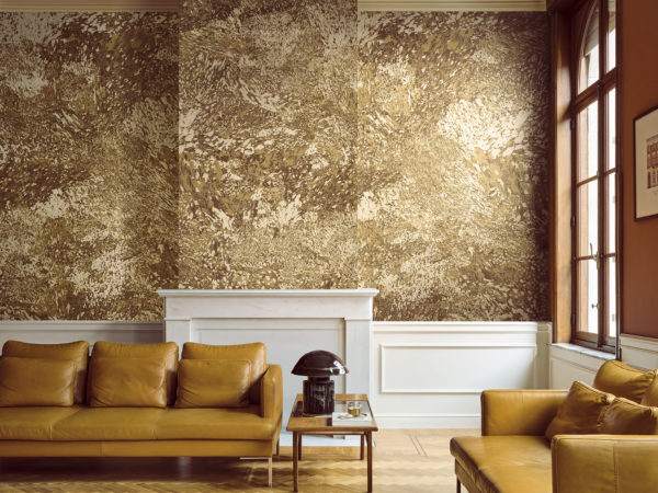 Elegantes Wohnzimmer mit der goldenen Whisper Wandbekleidung von Omexco, ein kontrastreicher Marmor mit malerischen Akzenten, veredelt durch einen sanft irisierenden Hintergrund. Die majestätische Wand umgibt einen klassischen weißen Kamin, ergänzt durch warme gelbe Ledersofas und einen Parkettboden.
